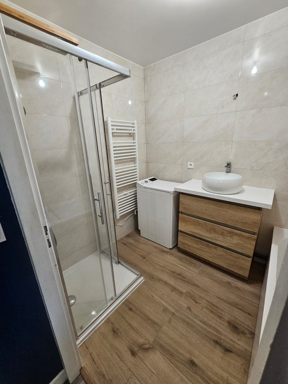 Salle de bain moderne avec douche vitrée, meuble en bois, vasque blanche et lave-linge blanc.