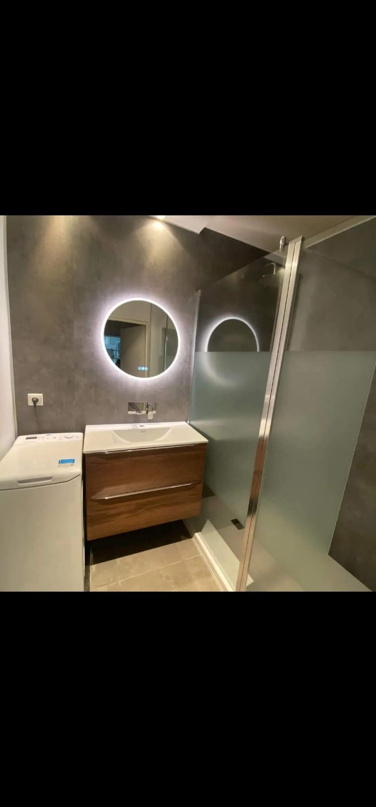Salle de bain moderne avec meuble vasque en bois, miroir rétroéclairé rond et douche vitrée.