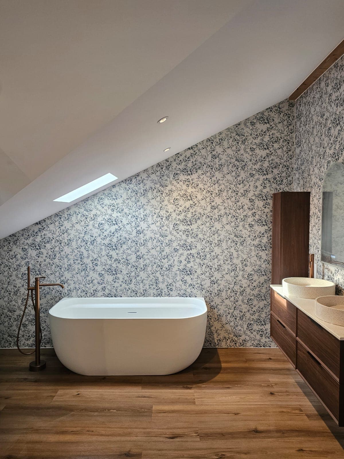 Baignoire blanche, papier peint floral bleu et meuble en bois sous un plafond mansardé.