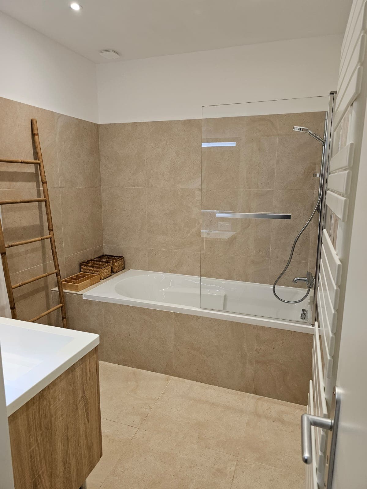 Salle de bain moderne avec carrelage beige, baignoire blanche, paroi vitrée et échelle en bambou.
