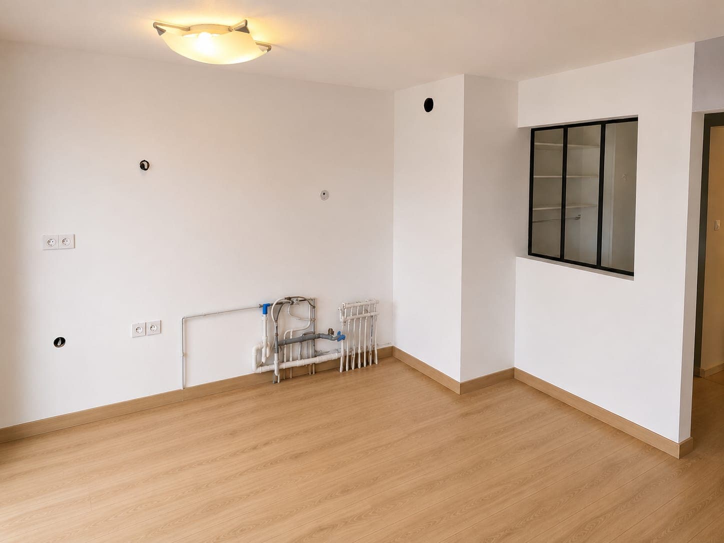 Pièce vide lumineuse avec murs blancs, parquet, tuyauterie apparente et petite verrière à cadre noir.