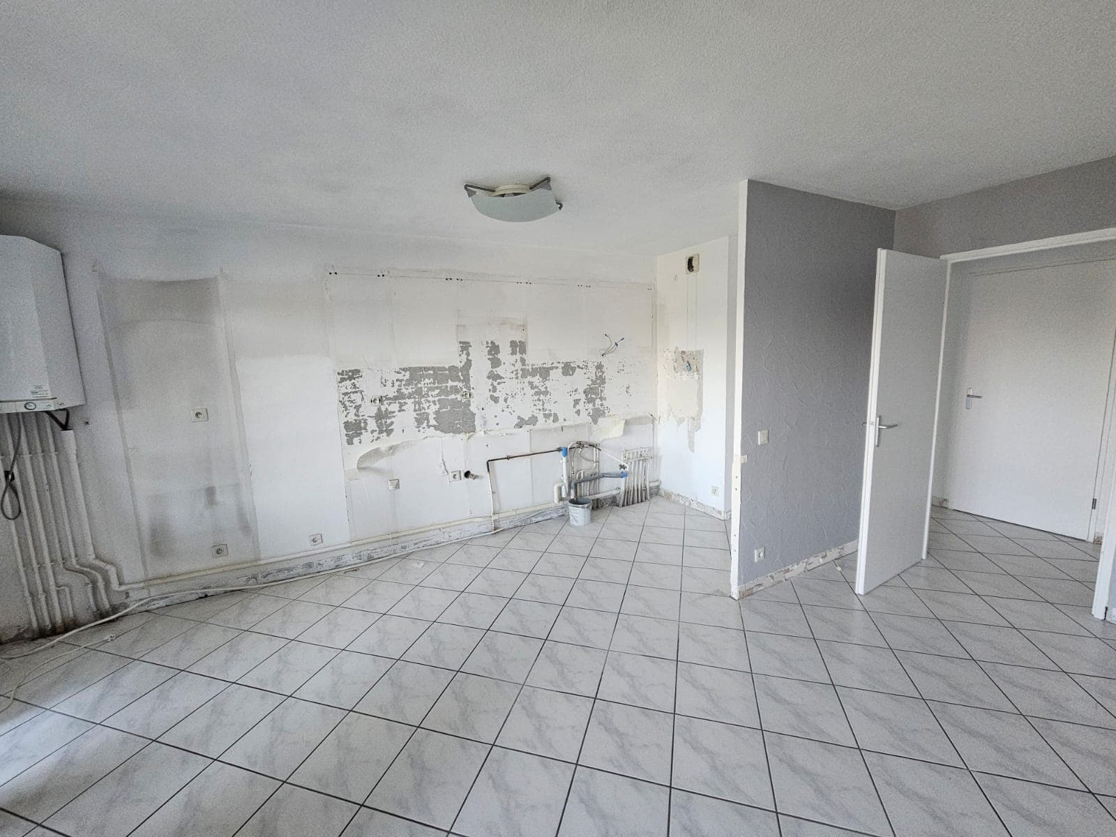 Pièce vide en rénovation avec carrelage blanc, murs abîmés et installations de plomberie apparentes.
